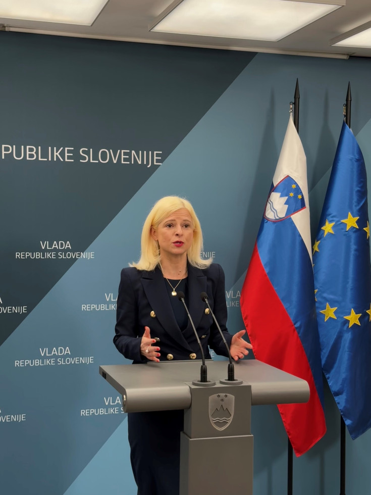 A blonde woman in a dark blazer is speaking at a podium during a press conference. Behind her are the flags of Slovenia and the European Union, along with a blue backdrop displaying Slovenian government branding.