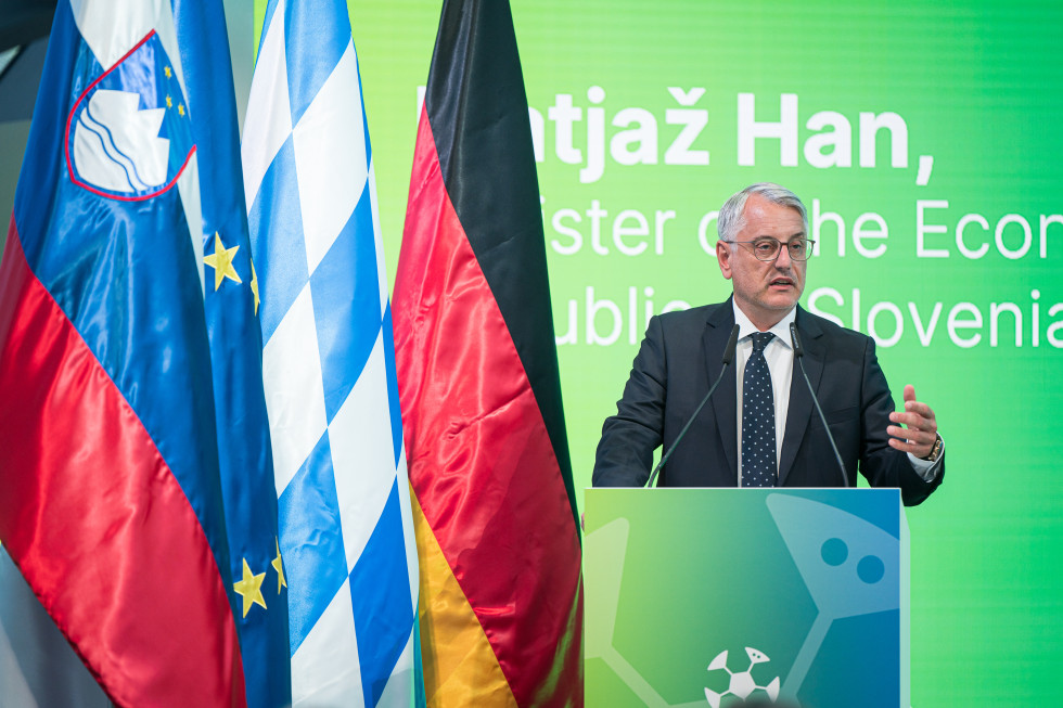 Minister Matjaž Han stoji za govorniškim pultom in nagovarja udeležence, za njim zelen zaslon, ob njem slovenska, evropska, bavarska in nemška zastava