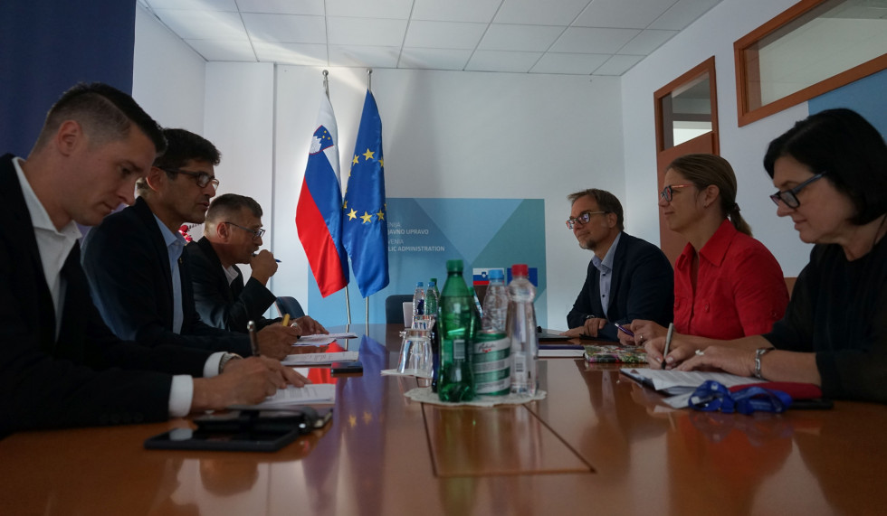 Sogovorniki sedijo v sejni sobi za ovalno mizo in se pogovarjajo. V ozadju slovenska in EU zastava.