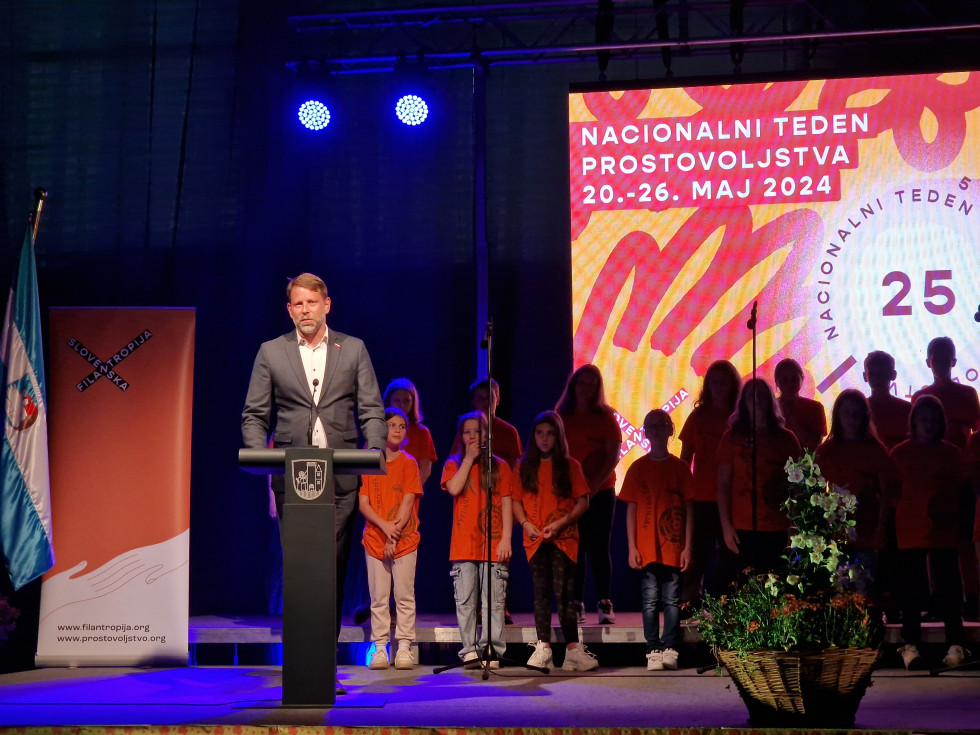 Za govornico stoji državni sekretar, levo od njega pano Slovenske filantropije, desno od njega stoji otroški pevski zbor. V ozadju je oranžen napis Nacionalni teden prostovoljstva. 