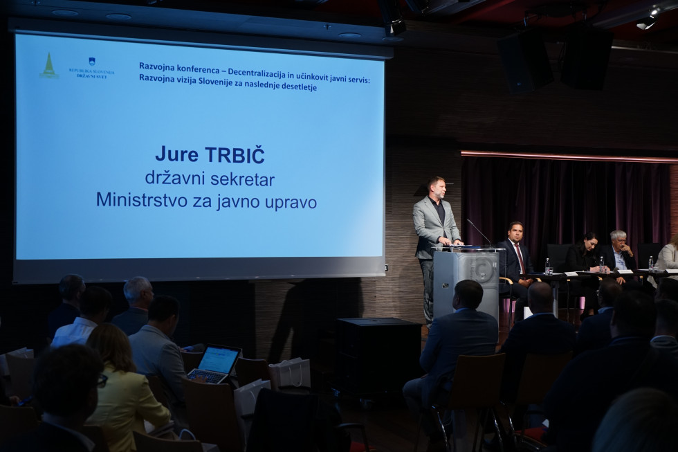 Državni sekretar Jure Trbič stoji pred govornico, v ozadju je napis razvojne konference Decentralizacija in učinkovit javni servis.