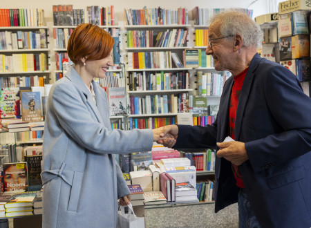 Minsitrica za kulturo se rokuje z moškim. Zadaj knjižna polica.