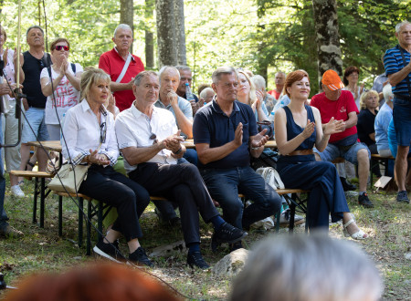 Ministrica za kulturo sedi na klopi med udeleženci spominske slovesnosti v Bazi 20 v Kočevskem rogu in ploska ob spremljanju slovesnosti. 