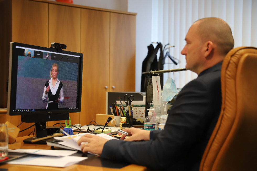 Moški v pisarni gleda v računalniški zaslon, na katerem je moderatorka konference med govorom.