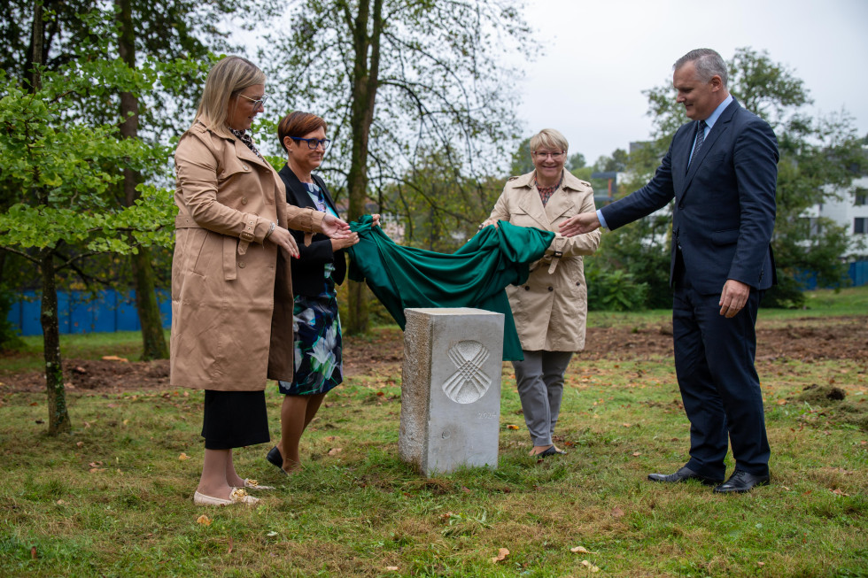 Več oseb stoji ob temeljnem kamnu in dviguje pregrinjalo. 