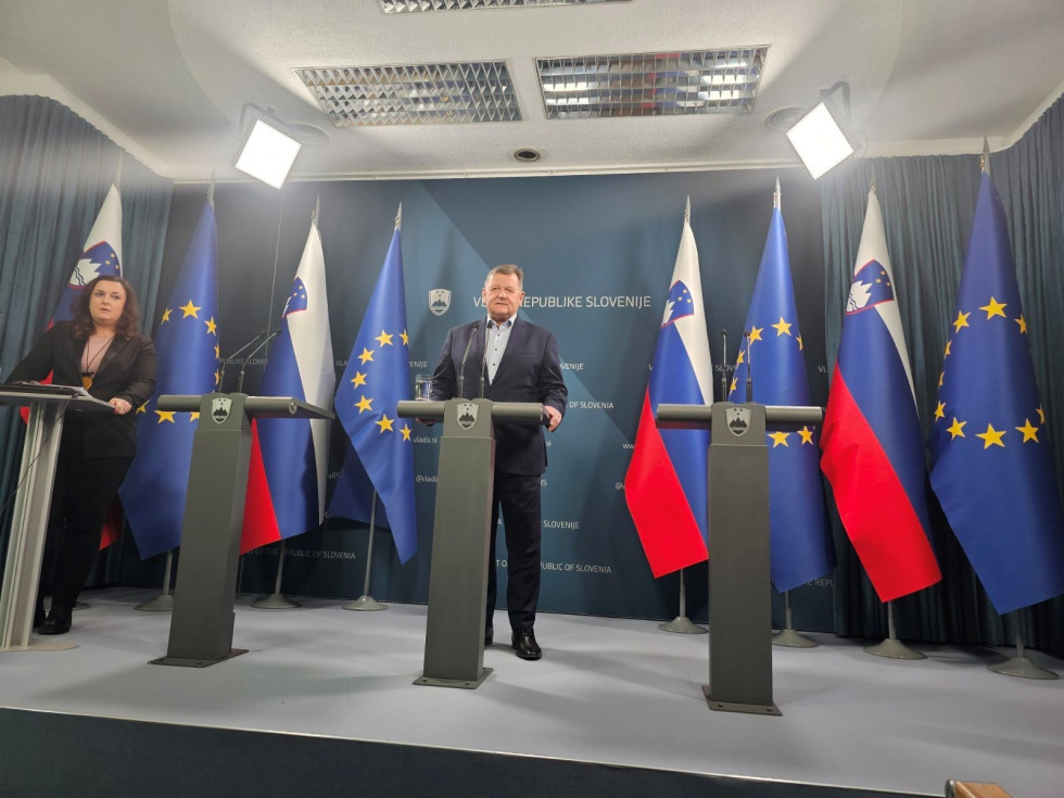 Minister Jevšek at the speaker's stand during a press conference at the Government Press Centre. Behind him are Slovenian and European flags.