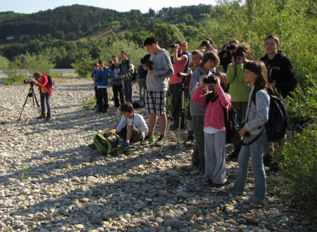 otroci in odrasli stojijo na brežini reke in opazujejo 