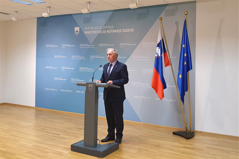Minister za notranje zadeve Aleš Hojs, stoji za govornico pred modrim panojem z napisom Ministrstvo za notranje zadeve. Desno sta zastavi Slovenije in Evropske unije.