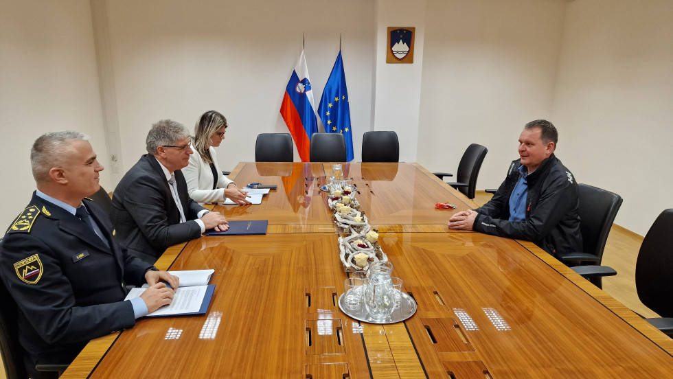 Ljudje sedijo na dveh straneh mize, pred njimi na rjavi mizi vrč z vodo in kozarci, v ozadju ob beli steni sta slovenska in evropska zastava