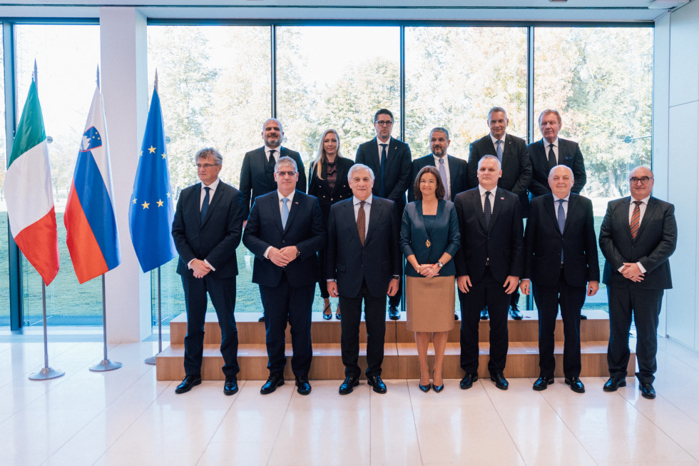 Ministers stand in two rows. Behind them are glass windows and the floor is light brown. On the left are the flags of Italy, Slovenia and the European Union.