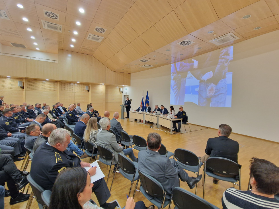 Udeleženci sedijo na stolih v več vrstah, pred njimi za katedrom sedijo panelisti, ob njih za govornico minister, za njim slovenska in evropska zastava, na beli steni v ozadju je fotografija policista, ki je vklenil osebo