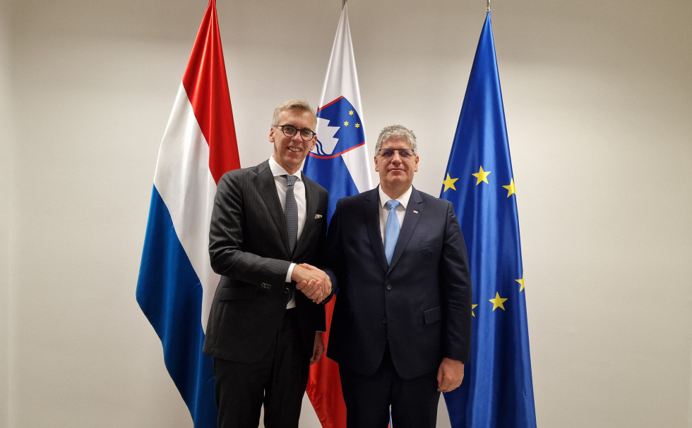 Ministers Boštjan Poklukar and Leon Gloden shake hands, with the Luxembourg, Slovenian and European flags behind them on the white wall