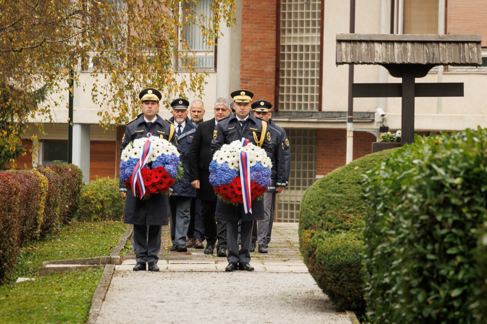 Udeleženci stojijo v dveh vrstah. Spredaj sta dva policista, vsak v svoji roki drži venec. Tla so makadamska, za njimi je bela stavba.