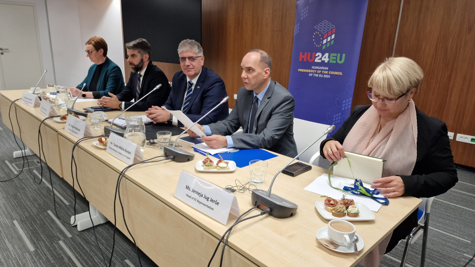 Trije moški in dve ženski sedijo za glavno mizo v notranjem prostoru. Na mizi so mikrofoni, vrči z vodo, kozarci, majhni sendviči, kava in tablice z njihovimi imeni. Za njimi je pokončen ožji pano z napisom HU24 EU.