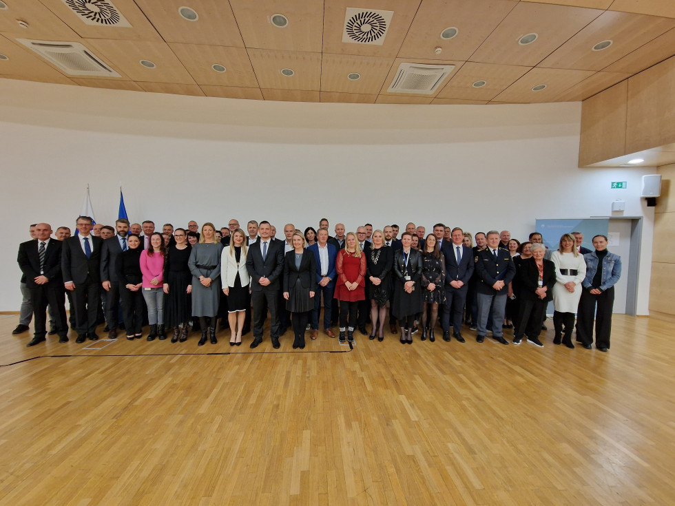 Udeleženci konference stojijo in se slikajo. Stena za njimi je bela, tla so svetlo rjave barve.