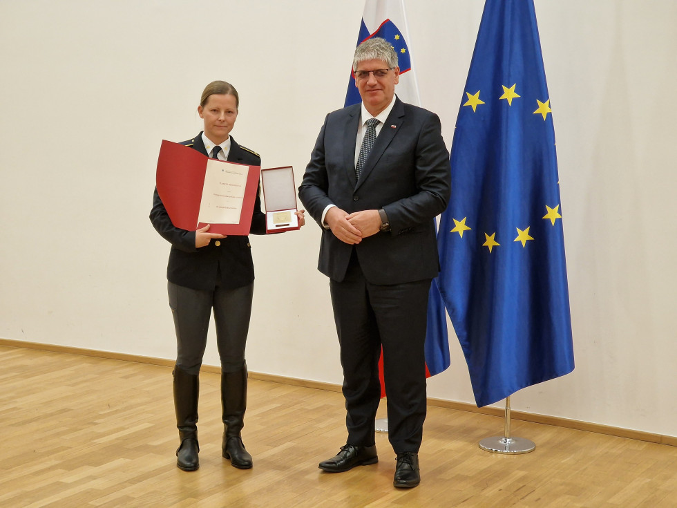 Minister in pomočnica načelnika stojita pred slovensko in evropsko zastavo, ki je ob beli steni