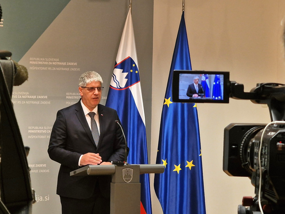 Il Ministro è in piedi dietro il leggio, con lo sguardo rivolto alle telecamere di fronte a lui, con la bandiera slovena e quella europea alle spalle.