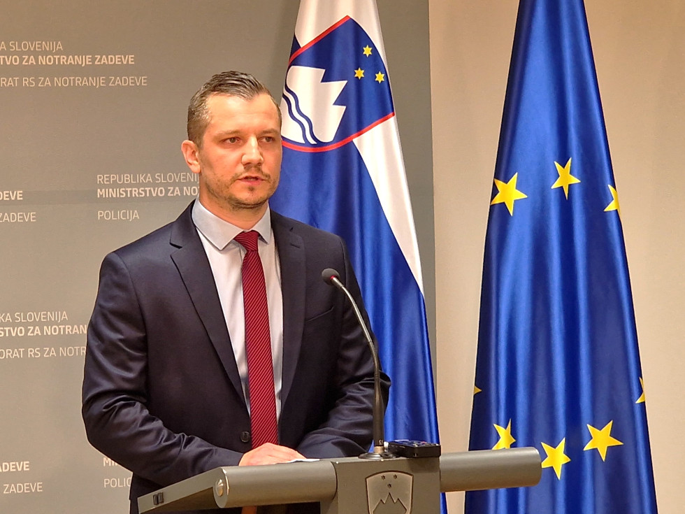 dr. Darijo Levačić stoji v notranjem prostoru za sivo govornico. Na desni sta slovenska in evropska zastava.