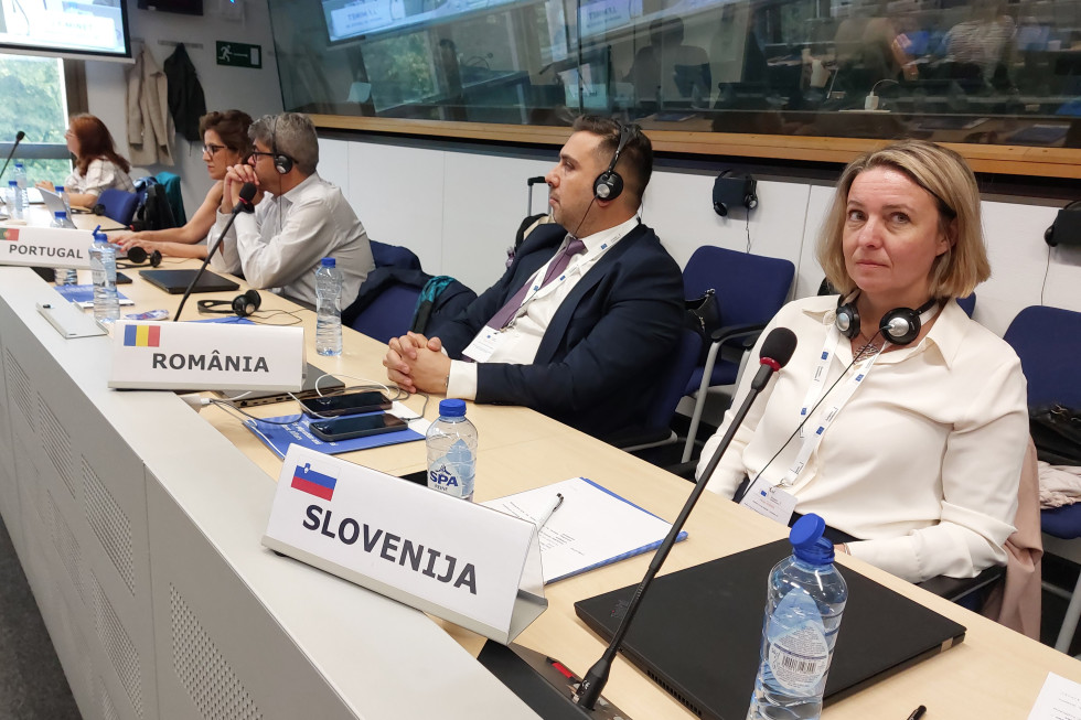 Državna sekretarka Helga Dobrin sedi za daljšo svetlo mizo. Pred njo je tablica z napisom Slovenija, desno od nje sedi predstavnik Romunije, na njegovi desni gospod iz Portugalske v svetlem. Dobrin nosi belo srajco, okrog vratu ima črne slušalke.