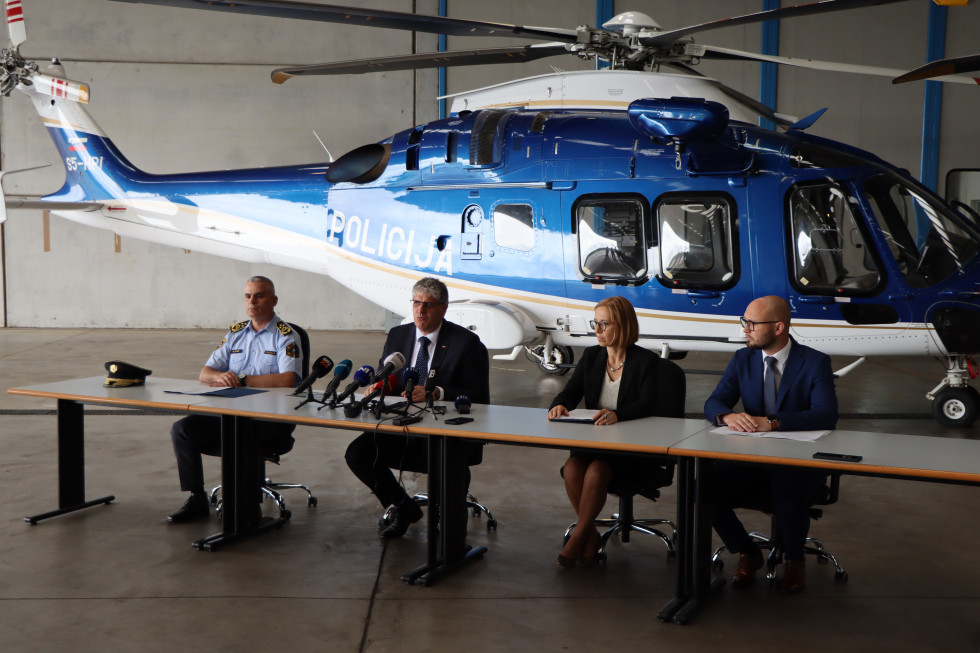 Govorci med novinarsko konferenco sedijo za mizo, na njej so postavljeni mikrofoni, za njimi stoji belo-moder policijski helikopter