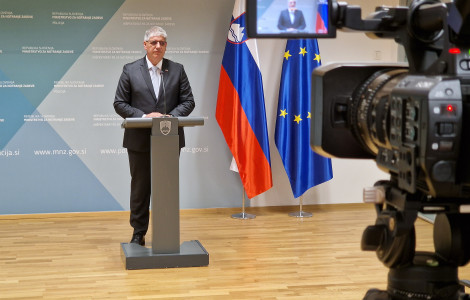 Izjava ministra za notranje zadeve Boštjana Poklukarja (Minister of the Interior Boštjan Poklukar stands behind the lectern in a bright room. Behind him is a blue billboard, with the Slovenian and European flags on the right. A camera with a screen and a picture of the Minister is visible.)