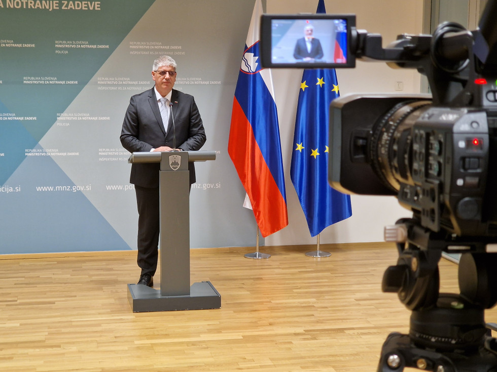 Minister of the Interior Boštjan Poklukar stands behind the lectern in a bright room. Behind him is a blue billboard, with the Slovenian and European flags on the right. A camera with a screen and a picture of the Minister is visible.