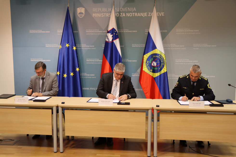 Tri osebe sedijo za bež mizami, na mizi so beli papirji, ki jih vsi trije podpisujejo. Za njimi modro ozadje, postavljene so tri zastave Evropske unije, Slovenije in Policije.