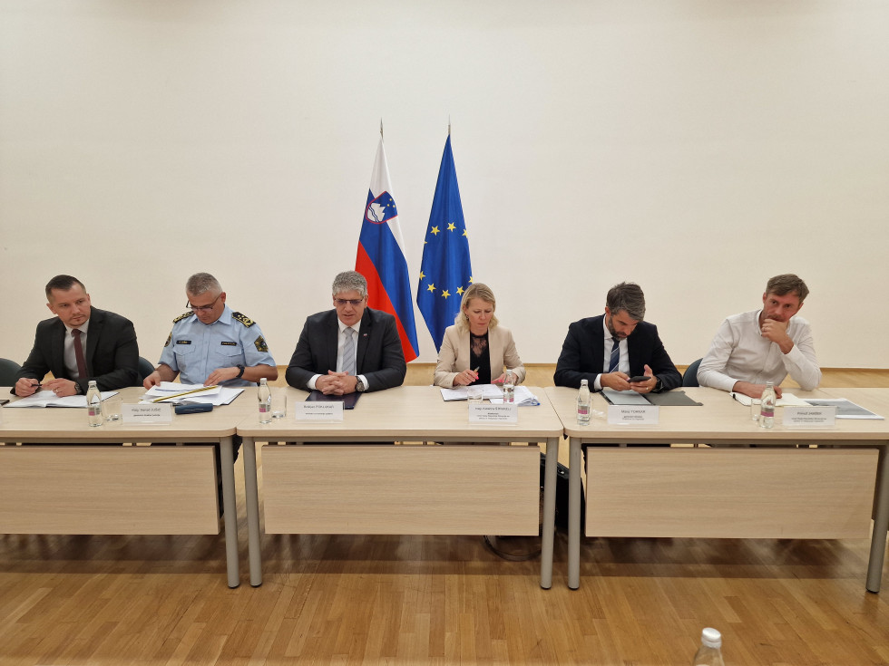 Predstavniki ministrstva in urada za oskrbo in integracijo migrantov sedijo za mizami bež barve. Za njimi zastava Sloveniije in Evropske unije, stena je bela, tla so svetlo rjave barve.
