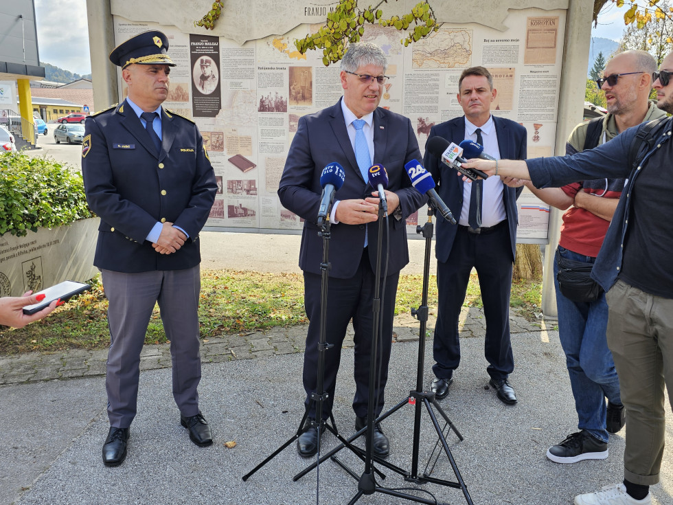 Minister Boštjan Poklukar stoji pred mikrofoni, na njegovi levi stoji generalni direktor policije mag. Senad Jušić, na desni pa župan Občine Šentjur Marko Diaci, skrajno desno stojijo novinarji, v ozadju so postavljene table z razstavo.