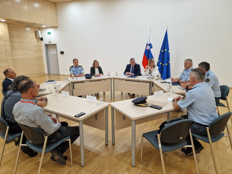 Srečanje vodstva ministrstva in policije s predstavniki romskih organizacij. Sedijo za mizami, ki so postavljene v krogu. Mize so svetlo rjave, dvorana je svetla, na stropu so majhne lučke, na tleh je svetel parket, za njimi je bela stena z zastavama.