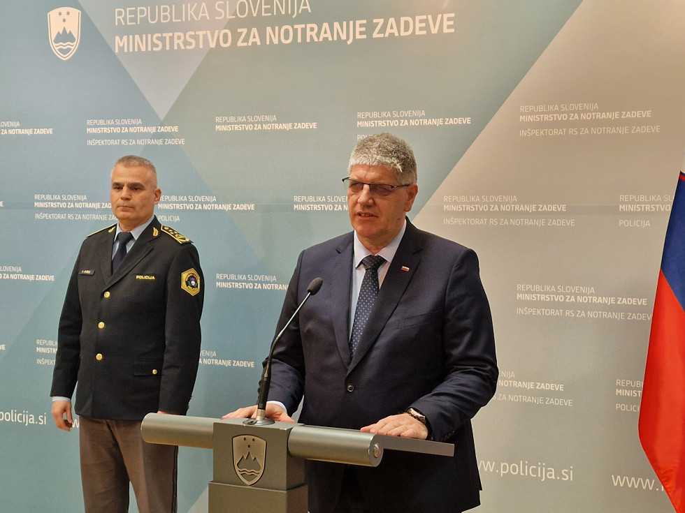 Minister stoji za govornico sive barve in govori, ob njem ob modri steni stoji generalni direktor policije