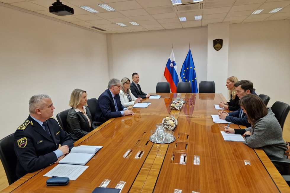 Več oseb sedi za svetlo rjavo lakirano sejno mizo. Stene in strop so beli, na stropu so vidne luči. Na mizi so vrči z vodo in kozarci. V ozadju sta slovenska in evropska zastava.