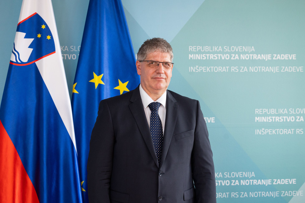 Minister Boštjan Poklukar stoji pred zastavama Slovenije in Evropske unije. Ozadje je v modrih barvah. Minister je oblečen v črno obleko, z belo srajco in črno kravato.