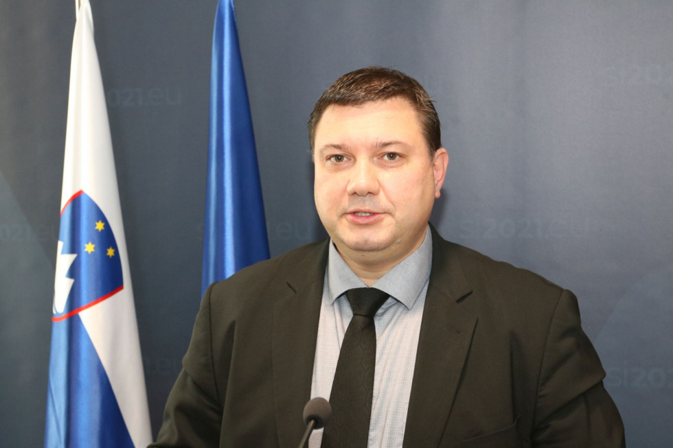 Direktor za govornico pred panojem predsedovanja in zastavama Slovenije in EU.