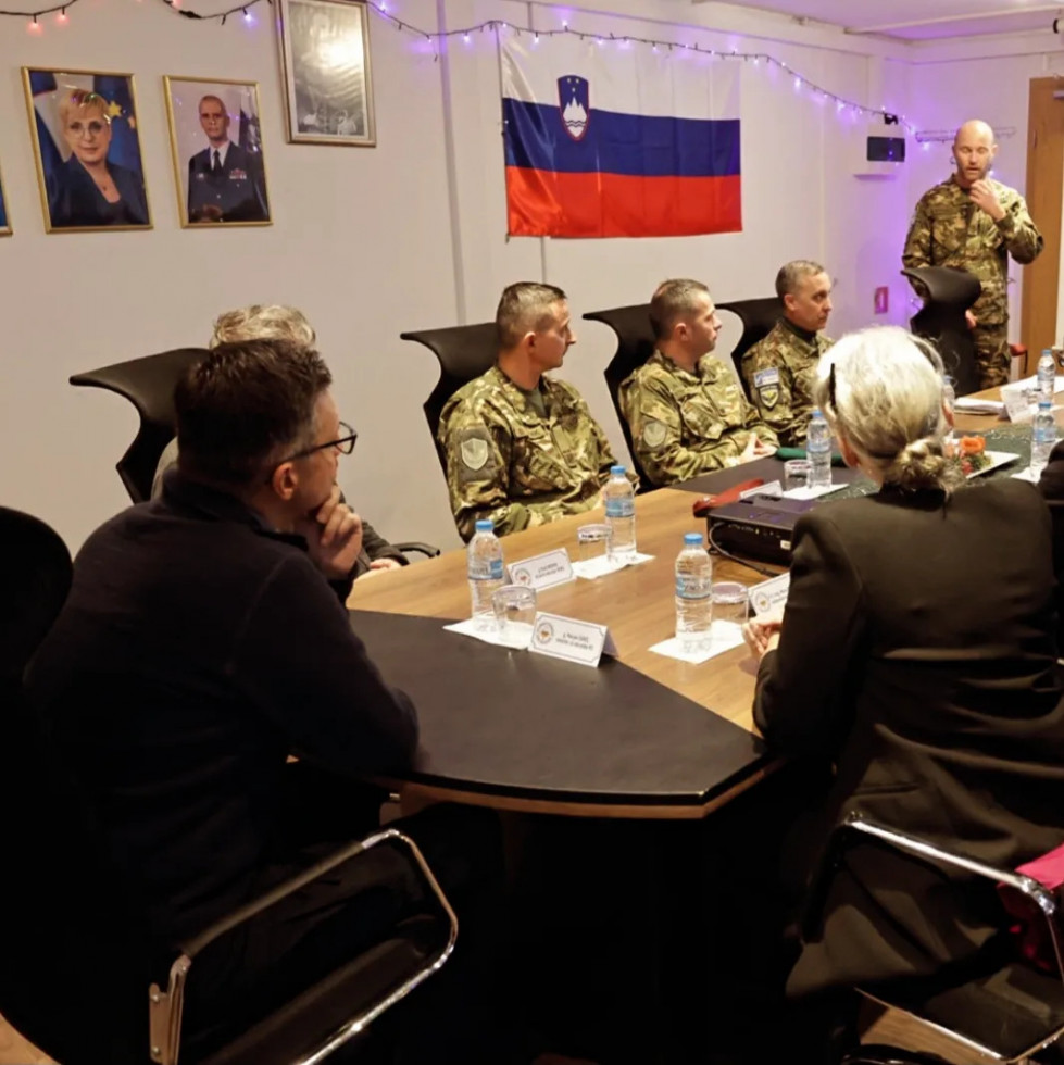 Minister sedi za mizo s predstavniki slovenskega kontingenta. Zadaj na steni je slovenska zastava