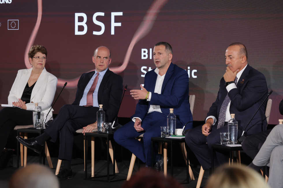 State Secretary Črnčec and other participating debaters are sitting on chairs on the stage, and in the background there is a white BSF sign on a dark billboard.