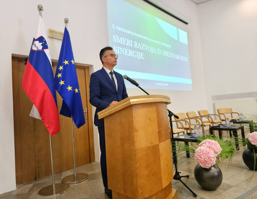 Minister med nastopom stoji za govornico. Za njim sta zastavi Slovenije in EU, ter platno s projekcijo
