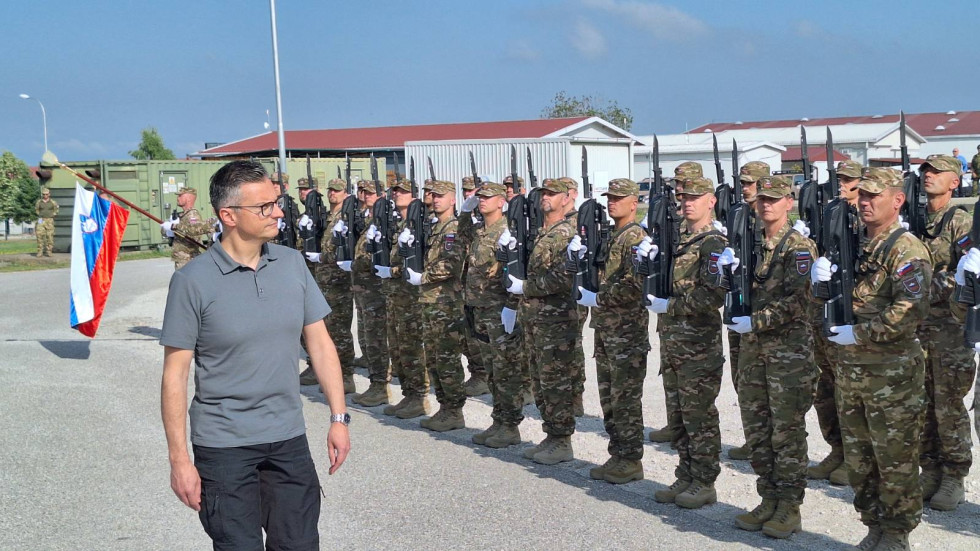 Minister Šarec stoji pred postrojem slovenskih vojakov, za njim je viden prapor s slovensko zastavo, ki ga drži v rokah vojak iz postroja, v ozadju so vojašnice baze v Novem Selu.