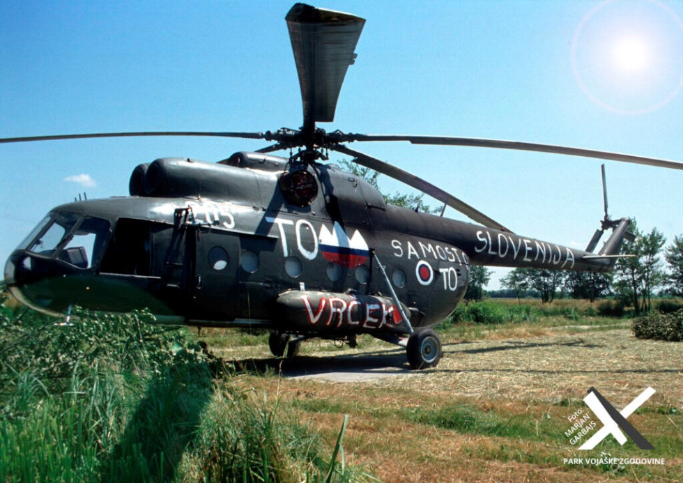 Helikopter stoji na travniku, na vojaško zeleni osnovni barvi so na levem boku bel napis TO s slovenskim grbom v barvah zastave, napis Samostojna TO in pod njima napis Vrček, na repu helikopterja pa bel napis Slovenija.
