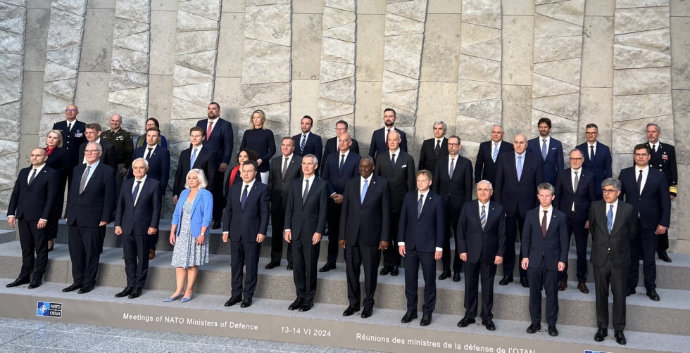 Ministri stojijo v treh vrstah na treh stopnicah, na prvi stopnici so angleški in francoski napis uradnega naziva srečanja ter znak Nata