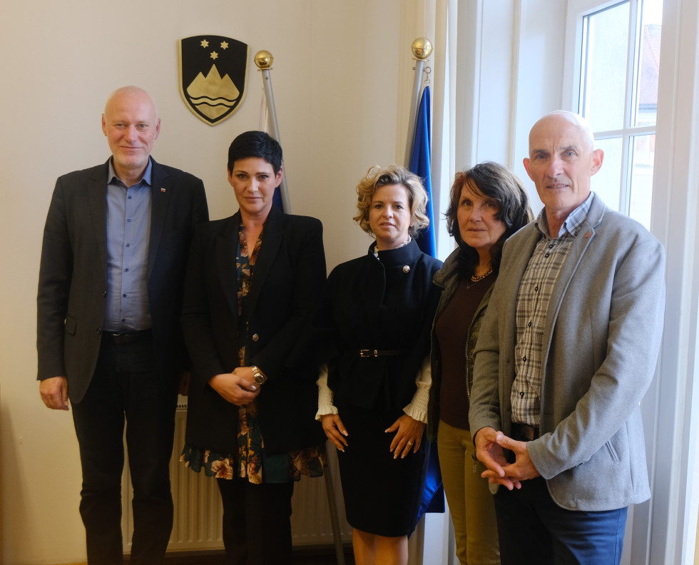 Skupinska fotografija državnega sekretarja dr. Milana Brgleza s predstavniki Društva sodnikov porotnikov. V ozadju slovenska in evropska zastava ter grb Slovenije.