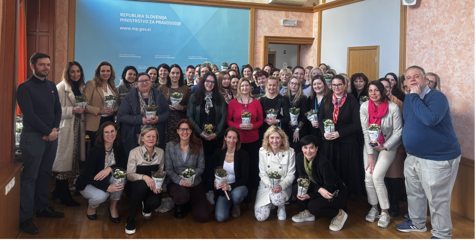 Skupinska fotografija nasmejanih sodelavk in sodelavcev z ministrico Andrejo Katič. Sodelavke so ob 8. marcu prejele rože. V ozadju tabla Ministrstva za pravosodje