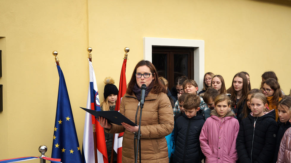 Tanja Malič stoji pred mikrofonom in nagovarja zbrane na odprtju, za njo članice in člani osnovnošolskega pevskega zbora