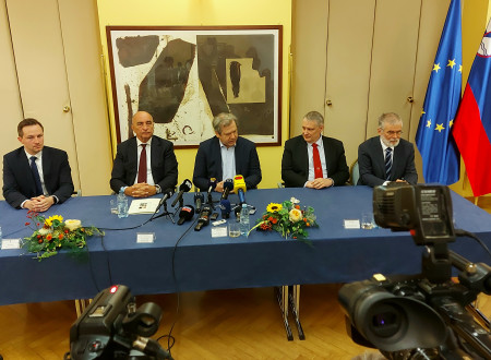 V ospredju dve kameri, v ozadju za mizo sedijo govorci med novinarsko konferenco. 