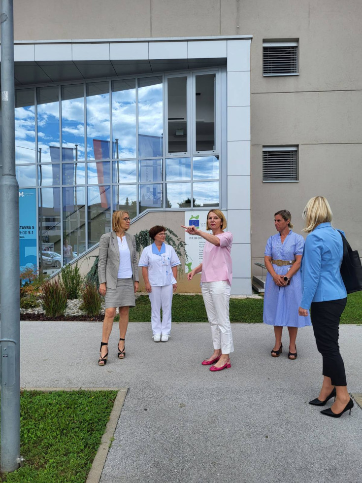 Pred steklenim vhodom v objekt stoji pet žensk. Ena je oblečena v belo zdravniško uniformo. Ženska na sredini ostalim nekaj kaže s prstom.