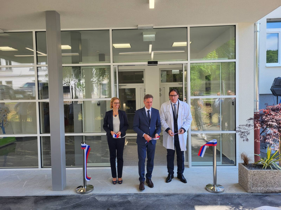 Ministrica, premier in direktor stojijo pred steklenim vhodom v stavbo. V rokah držijo del traku v barvah slovenske zastave. Ob straneh sta vidna kovinska stebrička, ki sta držala trak.