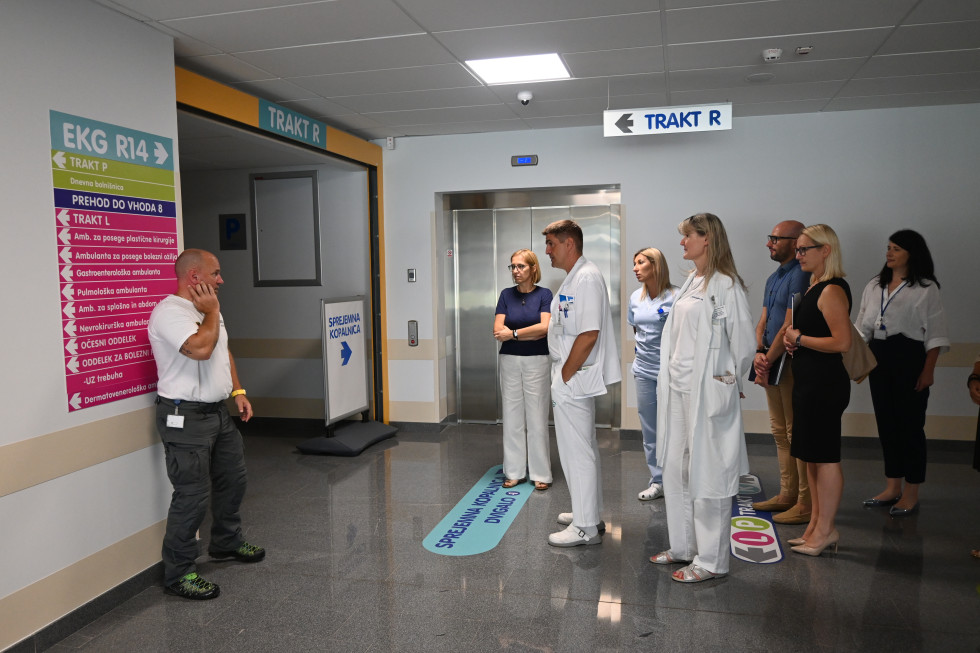 Na bolnišničnem hodniku stoji skupina ljudi, nekateri so v zdravniških haljah. Stojijo pred dvigalom in vhodom v naslednji hodnik. Ob vhodu stoji varnostnik. Na steni je usmeritvena tabla za različne oddelke.