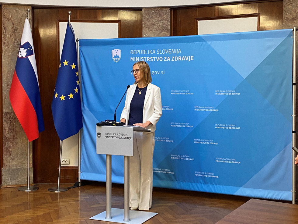 Ministrica za zdravje stoji pred modrim panojem, v levem kotu sta slovenska zastava in zastava Evropske unije. Stoji za govorniškim pultom. Oblečena je v bel kostim in modro majico.