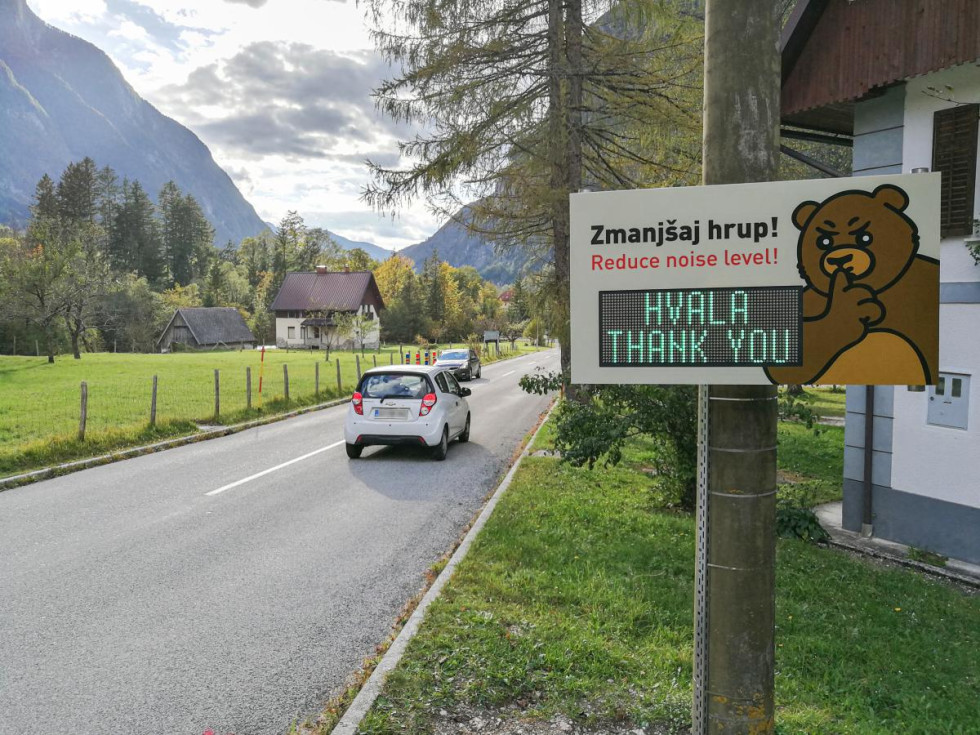 Cesta, na desni tabla z LED prikazovalnikom, ilustracija pedveda, s prstom čez "usta" simbolizira "Pssst". Na led zaslonu napis "Hvala. Thank you". Na cesti vozila. V ozadju visoko hribovje.