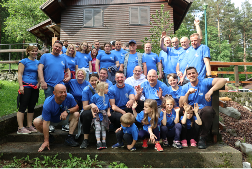 Udeleženci so vsi v modrih kratkih majicah, eni stojijo, drugi pa čepijo. V ozadju se vidi del del hiše.